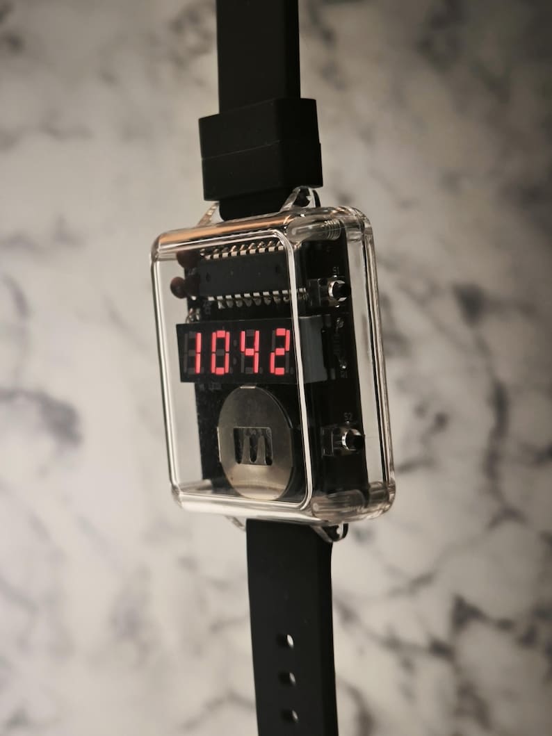 K&ouml;nnte beinhalten: Eine digitale Armbanduhr mit einem transparenten, rechteckigen Geh&auml;use, das die Uhrzeit 10:42 in roten Ziffern anzeigt. Die Uhr hat ein schwarzes Armband und einen schwarzen Stecker. Die inneren Komponenten der Uhr sind sichtbar.