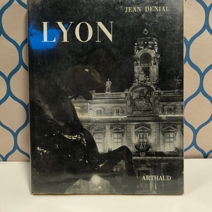 Può includere: Una fotografia in bianco e nero di un edificio a Lione, Francia. L'edificio ha una torre con un orologio ed è illuminato di notte. La copertina del libro riporta "LYON" e "JEAN DENIAU" e "ARTHAUD".