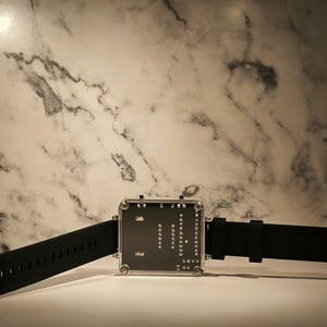 K&ouml;nnte beinhalten: Eine moderne Digitaluhr mit quadratischem Zifferblatt und schwarzem Silikonarmband. Das Zifferblatt zeigt ein Leiterplattendesign mit sichtbaren Komponenten. Die Uhr wird vor einem Marmorhintergrund pr&auml;sentiert.