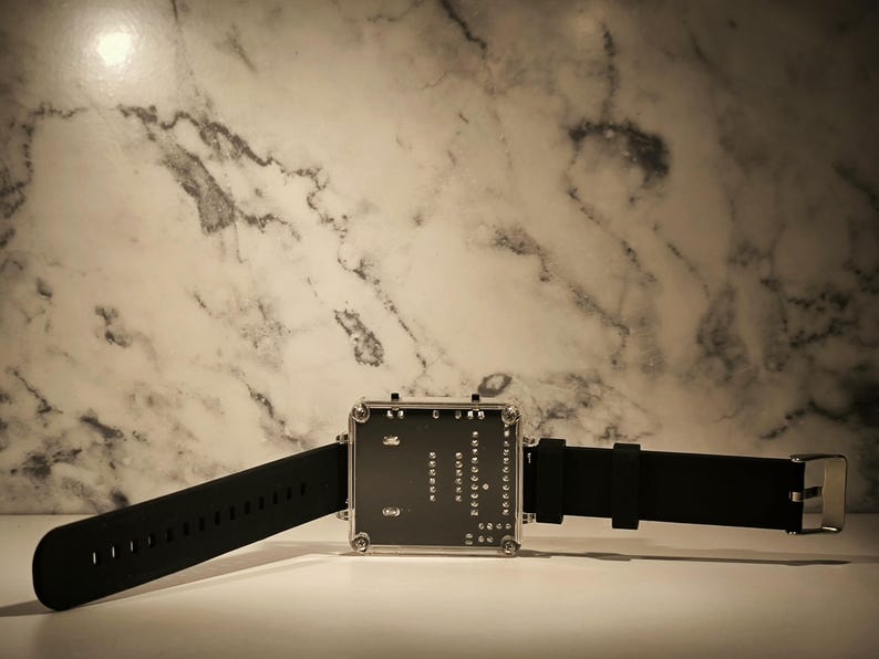 K&ouml;nnte beinhalten: Eine moderne Digitaluhr mit quadratischem Zifferblatt und schwarzem Silikonarmband. Das Zifferblatt zeigt ein Leiterplattendesign mit sichtbaren Komponenten. Die Uhr wird vor einem Marmorhintergrund pr&auml;sentiert.