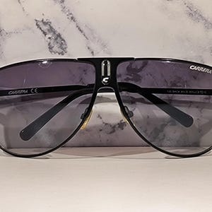 May include: Black Carrera sunglasses with a dark gray gradient lens. The sunglasses have a classic aviator shape with a black metal frame. The Carrera logo is visible on the top of the frame and on the lens.