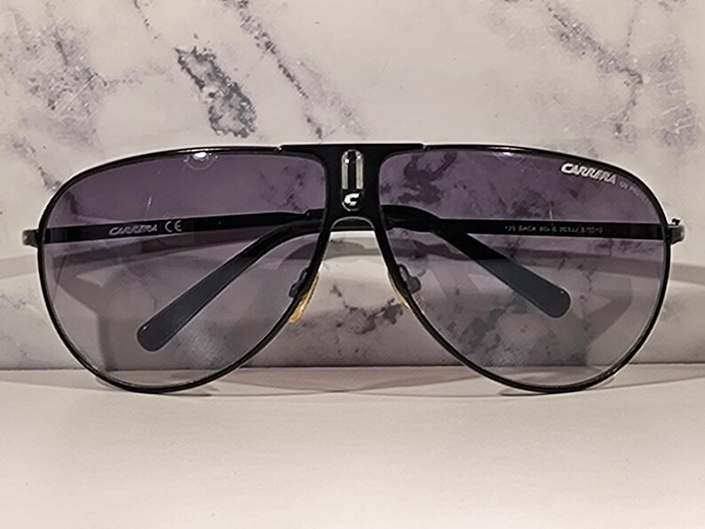 May include: Black Carrera sunglasses with a dark gray gradient lens. The sunglasses have a classic aviator shape with a black metal frame. The Carrera logo is visible on the top of the frame and on the lens.
