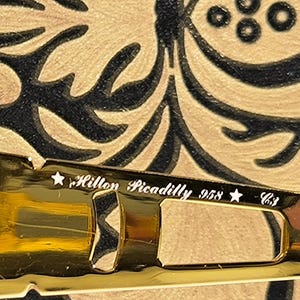 May include: Close-up of a gold-toned eyeglass frame with the text "Hilton Piccadilly 958 C3". The frame is set against a patterned background with black and gold swirls.
