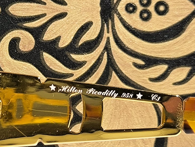 May include: Close-up of a gold-toned eyeglass frame with the text "Hilton Piccadilly 958 C3". The frame is set against a patterned background with black and gold swirls.