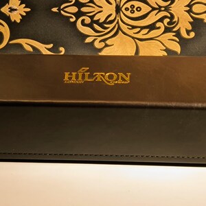 May include: A brown rectangular box with the gold text "HILTON LONDON eyewear" on the top. The box is made of a leather-like material and has a stitched edge. The background is a black and gold floral pattern.