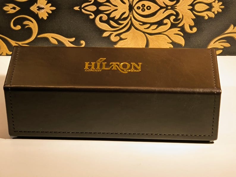 May include: A brown rectangular box with the gold text "HILTON LONDON eyewear" on the top. The box is made of a leather-like material and has a stitched edge. The background is a black and gold floral pattern.