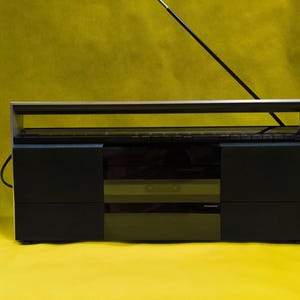 Può includere: Un impianto stereo vintage nero e argento con antenna retrattile. Lo stereo ha un design elegante e rettangolare con una cornice argentata e pannelli neri. Lo sfondo è giallo pieno.