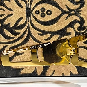 May include: Close-up of gold-colored sunglasses with a rectangular frame. The sunglasses have a gold hinge and the text "HILTON MADE IN ITALY CE 24KT" is visible on the arm. The background is a black and gold decorative pattern.