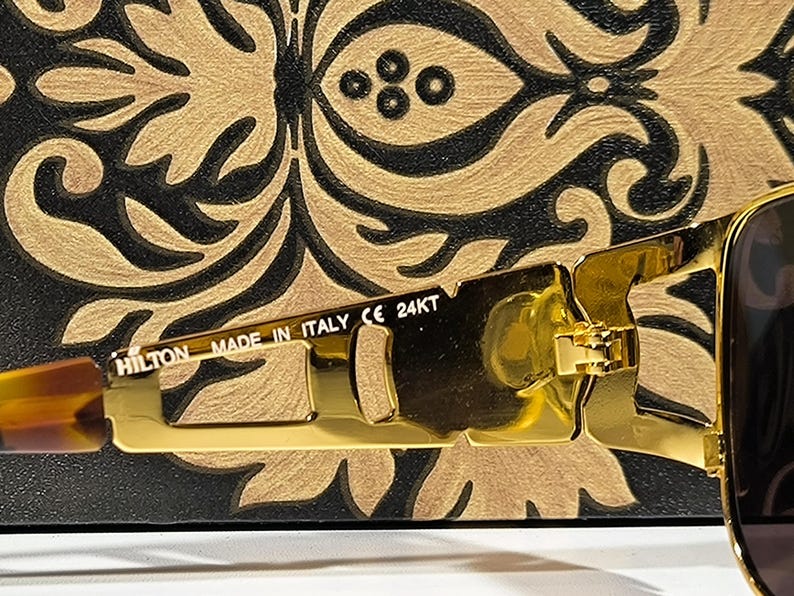 May include: Close-up of gold-colored sunglasses with a rectangular frame. The sunglasses have a gold hinge and the text "HILTON MADE IN ITALY CE 24KT" is visible on the arm. The background is a black and gold decorative pattern.