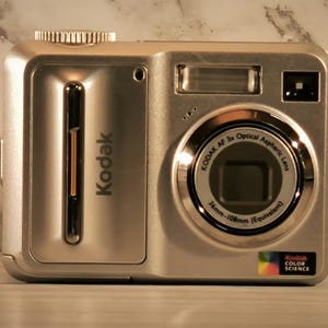 May include: Silver Kodak digital camera with a 3x optical zoom lens. The camera has a built-in flash and a color science logo. The lens has a 36mm-108mm equivalent focal length.