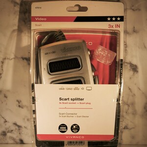 Könnte beinhalten: Ein Vivanco Scart-Splitter in einer transparenten Kunststoffverpackung. Die Verpackung hat rote und weiße Akzente und den Text "3x IN". Der Splitter ist silberfarben und hat mehrere Anschlüsse. Die Verpackung enthält auch den Text "Audio & Video".