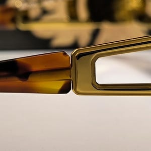 May include: Close-up of a pair of eyeglasses. The frame is gold-colored with a rectangular shape around the lenses. The arms of the glasses are a tortoiseshell pattern, with shades of brown and yellow. The glasses are on a white surface.