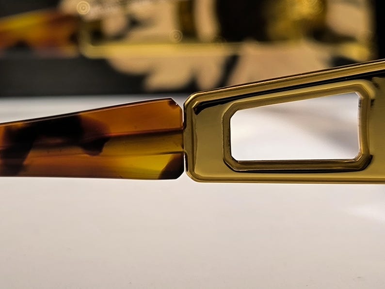 May include: Close-up of a pair of eyeglasses. The frame is gold-colored with a rectangular shape around the lenses. The arms of the glasses are a tortoiseshell pattern, with shades of brown and yellow. The glasses are on a white surface.