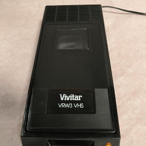 May include: Black Vivitar VRW3 VHS video player. The rectangular device has a clear window and the Vivitar logo with the model number. A power cord extends from the top. The device has a small, yellow light on the front.