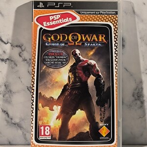 Pode incluir: Caixa de jogo PSP para "God of War: Ghost of Sparta". A capa apresenta um personagem musculoso e careca com marcas vermelhas, contra um fundo flamejante. A caixa inclui o texto "PSP Essentials" e "Uniquement sur PlayStation."