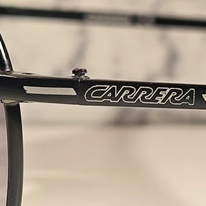 May include: Close-up of black Carrera sunglasses. The brand name "CARRERA" is displayed in white lettering on the temple arm. The sunglasses have a classic design with a dark lens and a black frame.