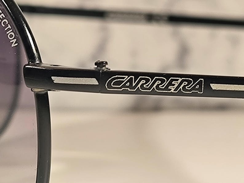 May include: Close-up of black Carrera sunglasses. The brand name "CARRERA" is displayed in white lettering on the temple arm. The sunglasses have a classic design with a dark lens and a black frame.