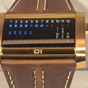 May include: A gold digital watch with a brown leather strap. The watch face shows the time and date with blue LED lights. The strap has white stitching and a rectangular gold case.