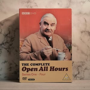 Pode incluir: Box de DVD de "The Complete Open All Hours: Series One - Four". A capa apresenta um homem com um casaco bege segurando uma colher e um pote. O logotipo da BBC DVD está no topo.