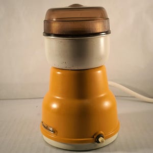 Può includere: Un macinacaffè elettrico vintage giallo e bianco con un coperchio in plastica trasparente. Il macinacaffè ha un cavo e un piccolo pulsante sul fondo.