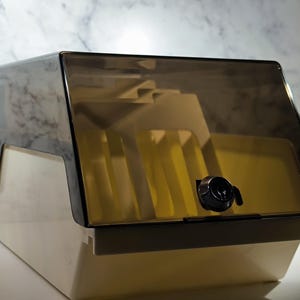 Peut inclure: Une boîte de rangement vintage, de couleur crème, avec un couvercle teinté marron et articulé. La boîte a une serrure noire et un intérieur jaune, probablement pour organiser des articles. Le couvercle est légèrement entrouvert, révélant le contenu.