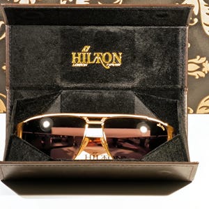 May include: Gold-framed sunglasses with brown lenses are displayed in a brown case. The case is open, revealing the sunglasses and a black interior. The brand name "HILTON LONDON" is visible in gold lettering.
