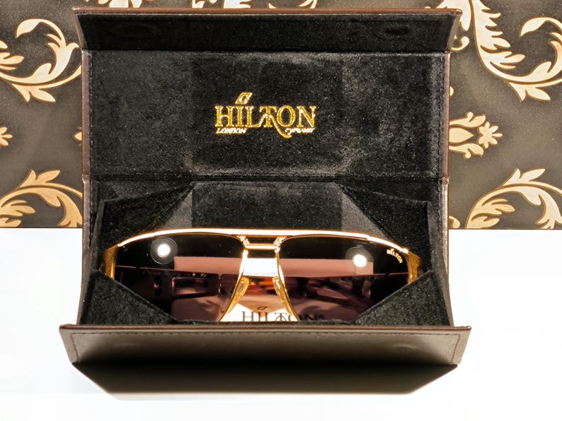 May include: Gold-framed sunglasses with brown lenses are displayed in a brown case. The case is open, revealing the sunglasses and a black interior. The brand name "HILTON LONDON" is visible in gold lettering.