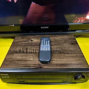 May include: A black Sharp VCR player with a wood grain effect top and a remote control. The VCR is on a yellow surface, with a Sony television in the background.