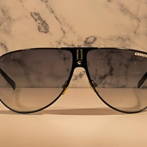 May include: Black Carrera sunglasses with a classic aviator design. The sunglasses have a black frame, gradient lenses, and the Carrera logo on the upper right lens. The lenses transition from dark at the top to lighter at the bottom.