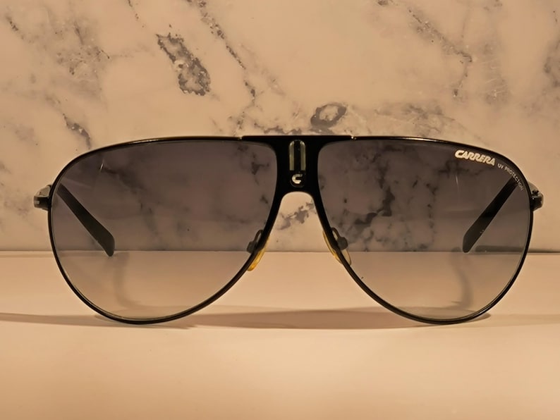 May include: Black Carrera sunglasses with a classic aviator design. The sunglasses have a black frame, gradient lenses, and the Carrera logo on the upper right lens. The lenses transition from dark at the top to lighter at the bottom.