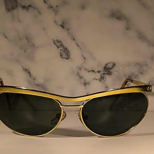 May include: Sunglasses with a polished gold-tone metal frame and dark green tinted lenses. The frame features a distinct brow bar design that connects the two rounded lenses, with thin gold-tone metal temples extending from the sides.