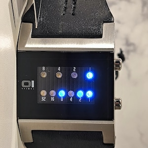 May include: A digital wristwatch with a black leather strap and a rectangular face. The watch face displays time using illuminated blue dots and numbers. The watch has a silver-coloured metal casing and the brand name "OI THEONE" is visible.