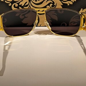 May include: Gold-framed sunglasses with dark lenses. The sunglasses have a double bridge and a rectangular shape. The temples are a tortoiseshell pattern. The sunglasses are set against a decorative background.