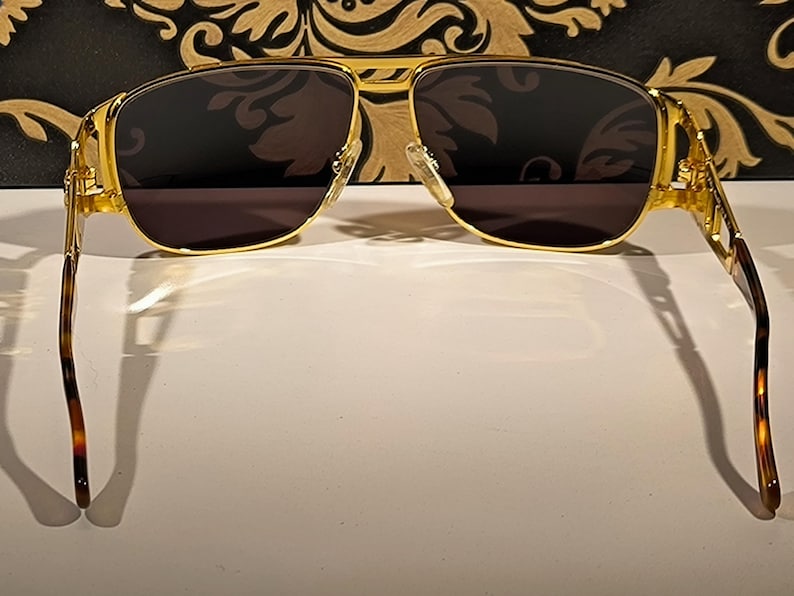 May include: Gold-framed sunglasses with dark lenses. The sunglasses have a double bridge and a rectangular shape. The temples are a tortoiseshell pattern. The sunglasses are set against a decorative background.
