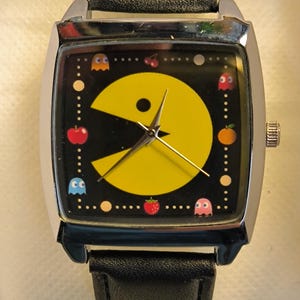 May include: A retro wristwatch with a black face displaying a yellow Pac-Man character, ghosts, and fruit. The watch has a silver case and a black leather strap. A nostalgic accessory, reminiscent of the classic arcade game.