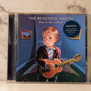 Puede incluir: Caja de CD del álbum "The Beautiful South", "blue is the colour". La portada muestra a un niño con una guitarra en una habitación azul. Una etiqueta de advertencia es visible. El CD está en una funda de plástico transparente.