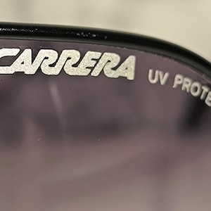 May include: Close-up of sunglasses with the brand name "CARRERA" in silver lettering. The text "UV PROTECTION" is also visible. The lenses are a dark purple color, and the frame is black.