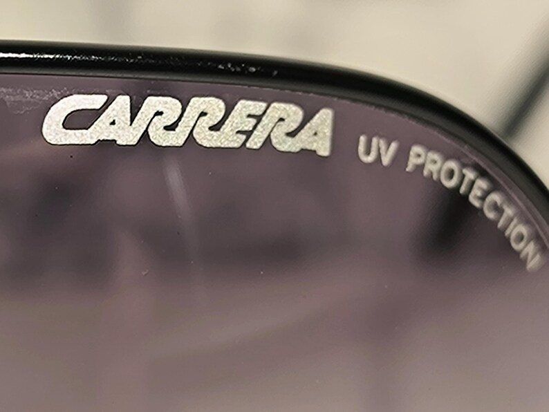 May include: Close-up of sunglasses with the brand name "CARRERA" in silver lettering. The text "UV PROTECTION" is also visible. The lenses are a dark purple color, and the frame is black.