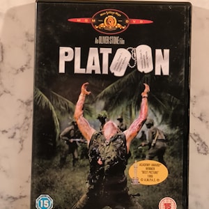 Puede incluir: Caja de DVD de la película "Platoon" de Oliver Stone. La portada presenta a un hombre con los brazos levantados, soldados al fondo y el título de la película. Incluye una pegatina de los Premios de la Academia y el texto "The first casualty of war is innocence."