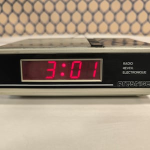 May include: A silver digital clock with a red display showing the time 3:01. The clock has the words "RADIO REVEIL ELECTRONIQUE" and the brand name "PRESTIGE" printed on it.