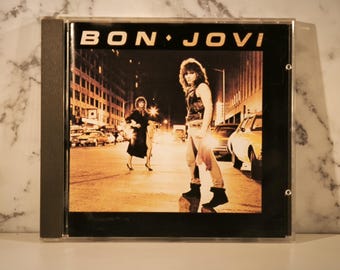 Puede incluir: Caja de CD con la portada del álbum de Bon Jovi. La imagen muestra a una persona caminando por una calle de la ciudad por la noche, con el nombre de la banda en la parte superior. La paleta de colores es cálida, con tonos naranja y marrón.