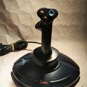 Puede incluir: Joystick negro de la marca Genius con base redonda y palanca central. El joystick tiene un cable negro y algunos botones de colores en la parte superior. La base tiene la palabra "Genius" en rojo.