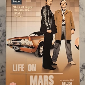 Könnte beinhalten: DVD-Hülle der britischen Fernsehserie "Life on Mars". Das Cover zeigt zwei Männer, ein Oldtimer und den Titel der Serie. Die Hülle ist braun mit dem BBC-Logo und einem Zitat von Radio Times. Die komplette erste Staffel.