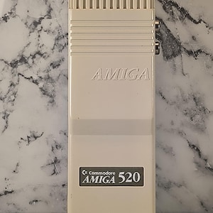 Puede incluir: Un adaptador de vídeo Commodore Amiga 520 beige vintage. El dispositivo tiene la palabra "AMIGA" en relieve en la parte delantera y una etiqueta que dice "Commodore AMIGA 520". La parte superior tiene crestas verticales y varios puertos.