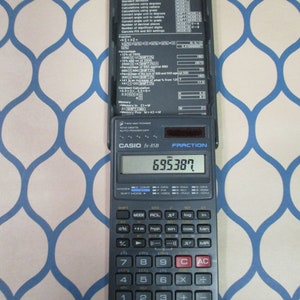 May include: A black Casio fx-85B scientific calculator with a silver display showing the number 695387. The calculator has a variety of buttons, including trigonometric functions, logarithms, and exponents. The calculator also has a fraction button and a memory button.