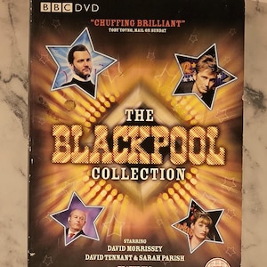 Może przedstawiać: Pudełko DVD dla "The Blackpool Collection" ze zdjęciami aktorów w ramkach w kształcie gwiazd. Okładka jest przeważnie czarna i złota, z tytułem dużymi, podświetlanymi literami. Logo BBC DVD znajduje się w lewym górnym rogu.