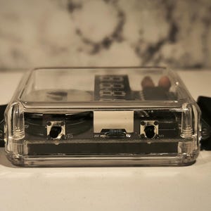 K&ouml;nnte beinhalten: Ein klarer, rechteckiger Uhrenkasten mit digitaler Zeitanzeige. Die Uhr hat ein schwarzes Armband und zwei Kn&ouml;pfe auf der Vorderseite. Das Geh&auml;use besteht aus transparentem Kunststoff.