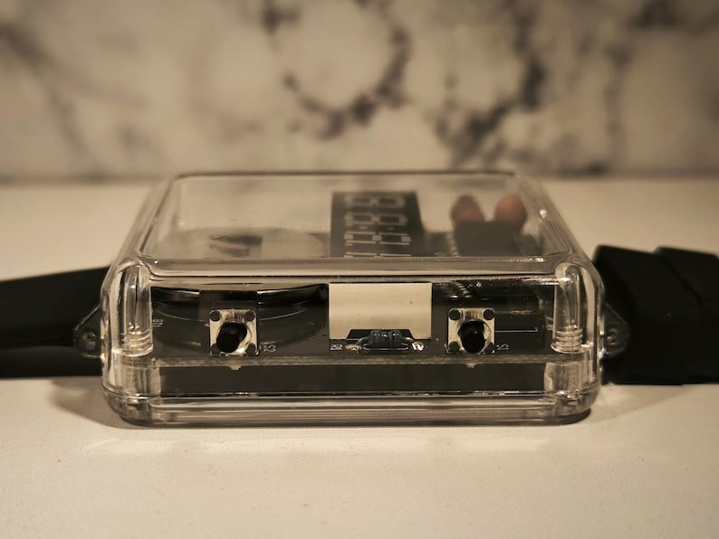 K&ouml;nnte beinhalten: Ein klarer, rechteckiger Uhrenkasten mit digitaler Zeitanzeige. Die Uhr hat ein schwarzes Armband und zwei Kn&ouml;pfe auf der Vorderseite. Das Geh&auml;use besteht aus transparentem Kunststoff.