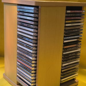 Op de afbeelding: Een licht houten cd-opbergkast gevuld met compact discs. De kast heeft een eenvoudig, rechthoekig ontwerp en is gemaakt van lichtgekleurd hout. De planken zijn vol cd's, met een muziekcollectie.