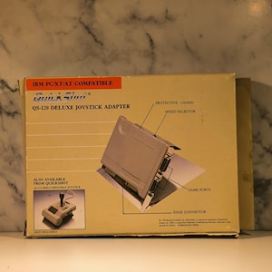 May include: A vintage QuickShot QS-120 Deluxe Joystick Adapter box. The box is beige with text and diagrams, including "IBM PC/XT/AT COMPATIBLE" and "QS-120 DELUXE JOYSTICK ADAPTER". The adapter is shown with a protective casing, speed selector, game ports, and edge connector.